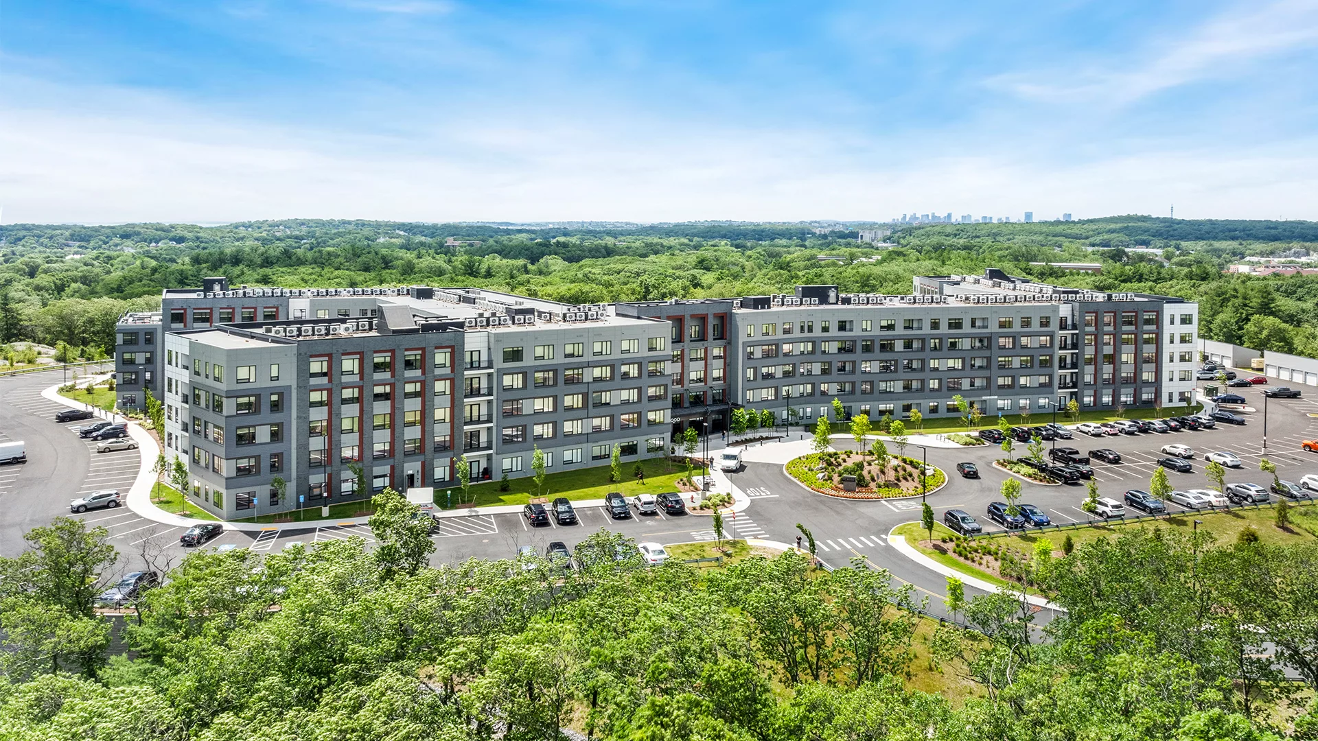 Drone photo of the exterior of a grey and white apartment building surrounded by a parking lot with green trees and a city skyline in the background.