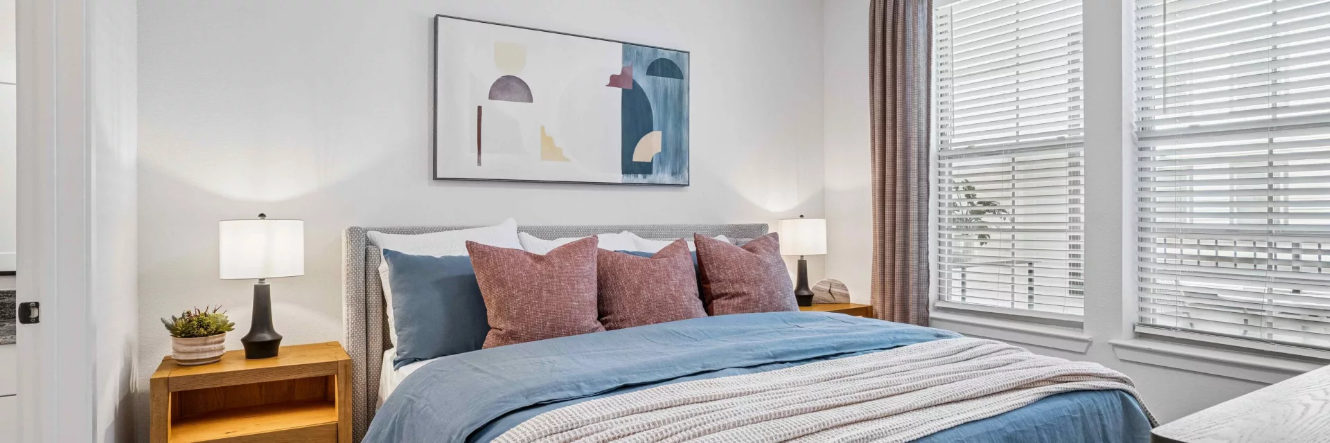 Model bedroom with blue bedding, nightstands, lamps, a ceiling fan, and windows with blinds.
