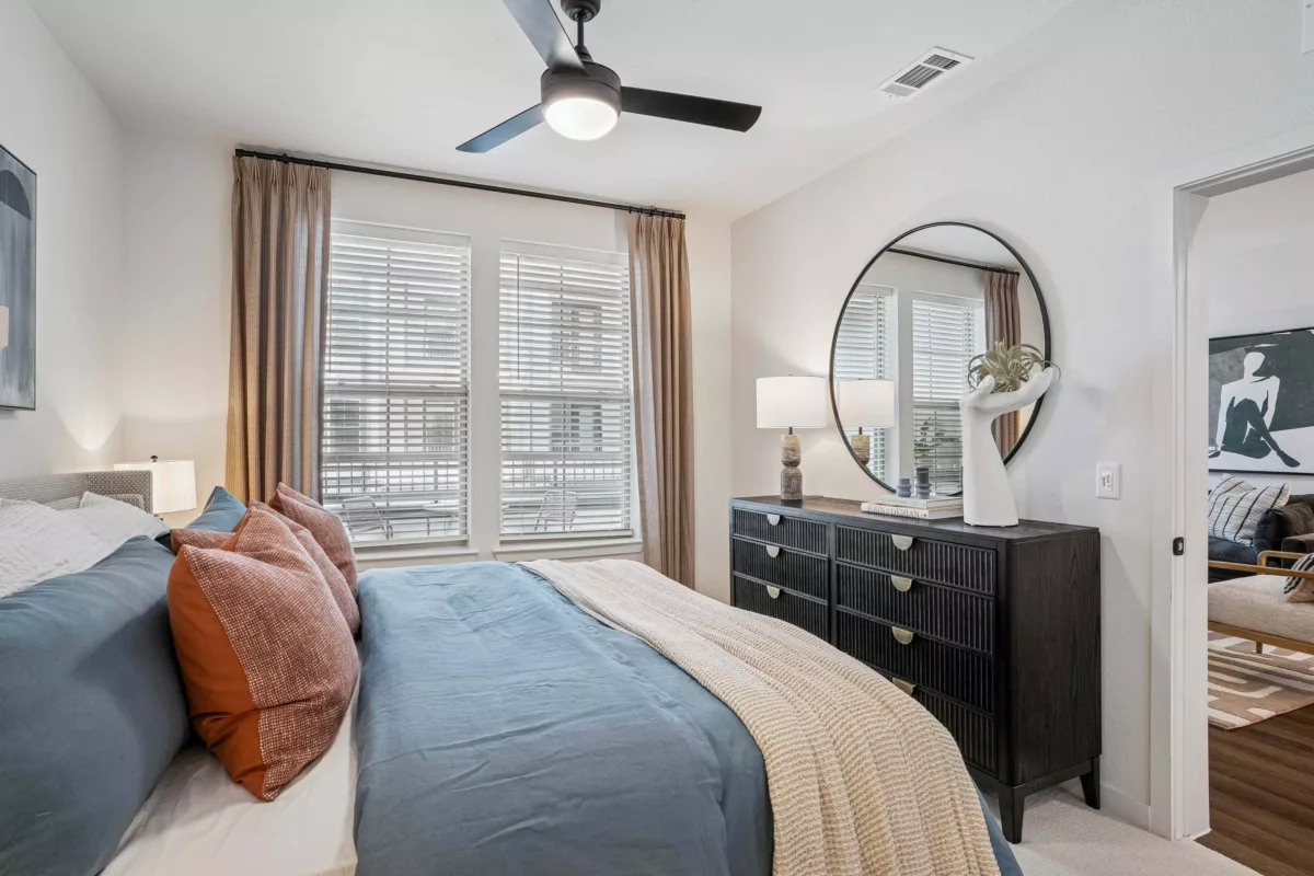 Model bedroom with blue bedding, dresser with round mirror, and window with blinds.