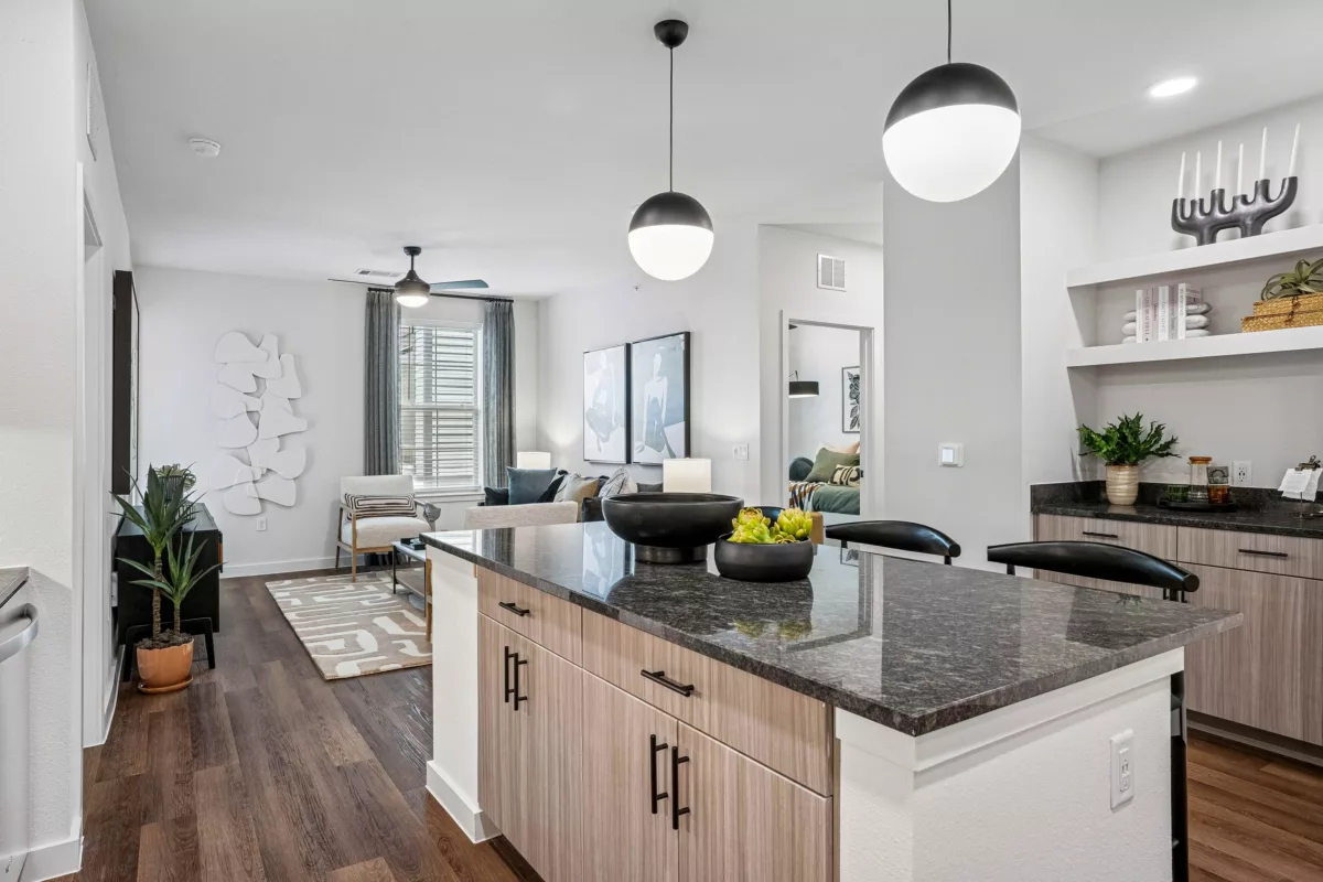 Open-concept kitchen with granite island, pendant lights, and wood-look cabinets, overlooking a living room with white walls and gray curtains.