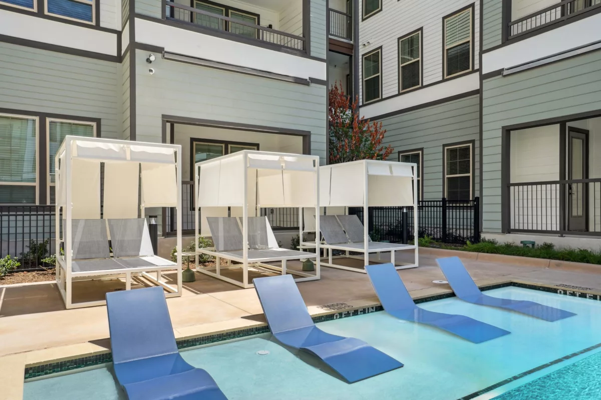 Poolside cabanas and blue lounge chairs in shallow water area. Building in background.