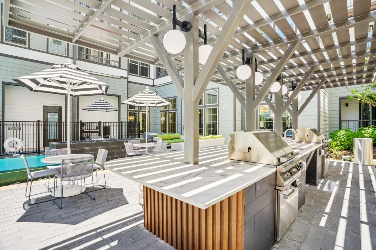 Outdoor kitchen with stainless steel gas grills under a pergola with globe lights. Patio seating nearby.