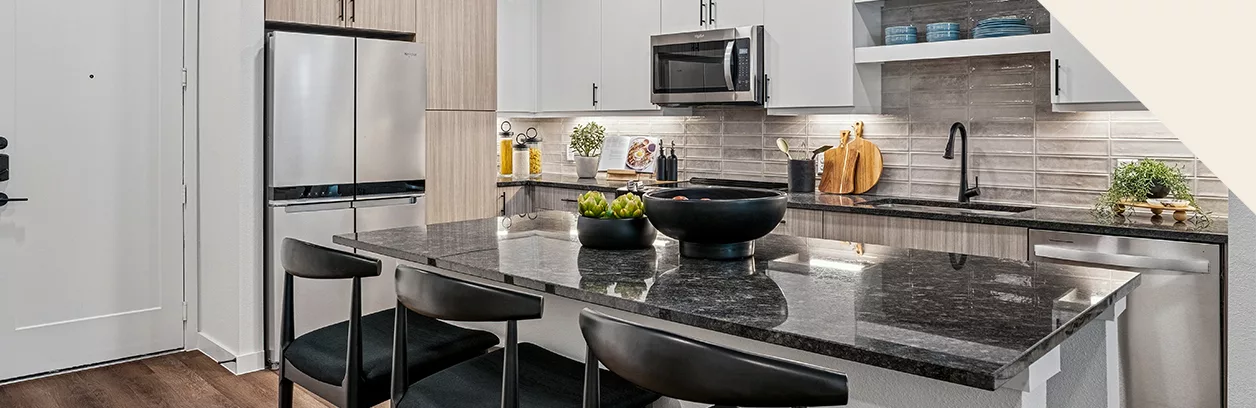 A kitchen with granite countertops, stainless steel appliances, white and light wood cabinets, and a island with black stools.