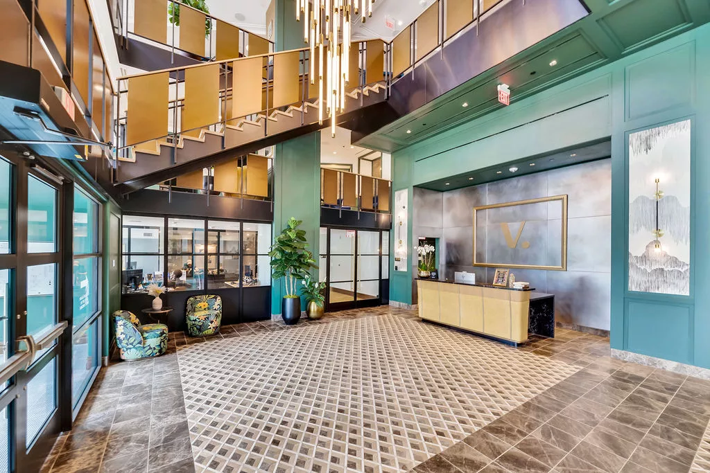Grand lobby with staircase, gold chandelier, and reception desk with “V.” logo.