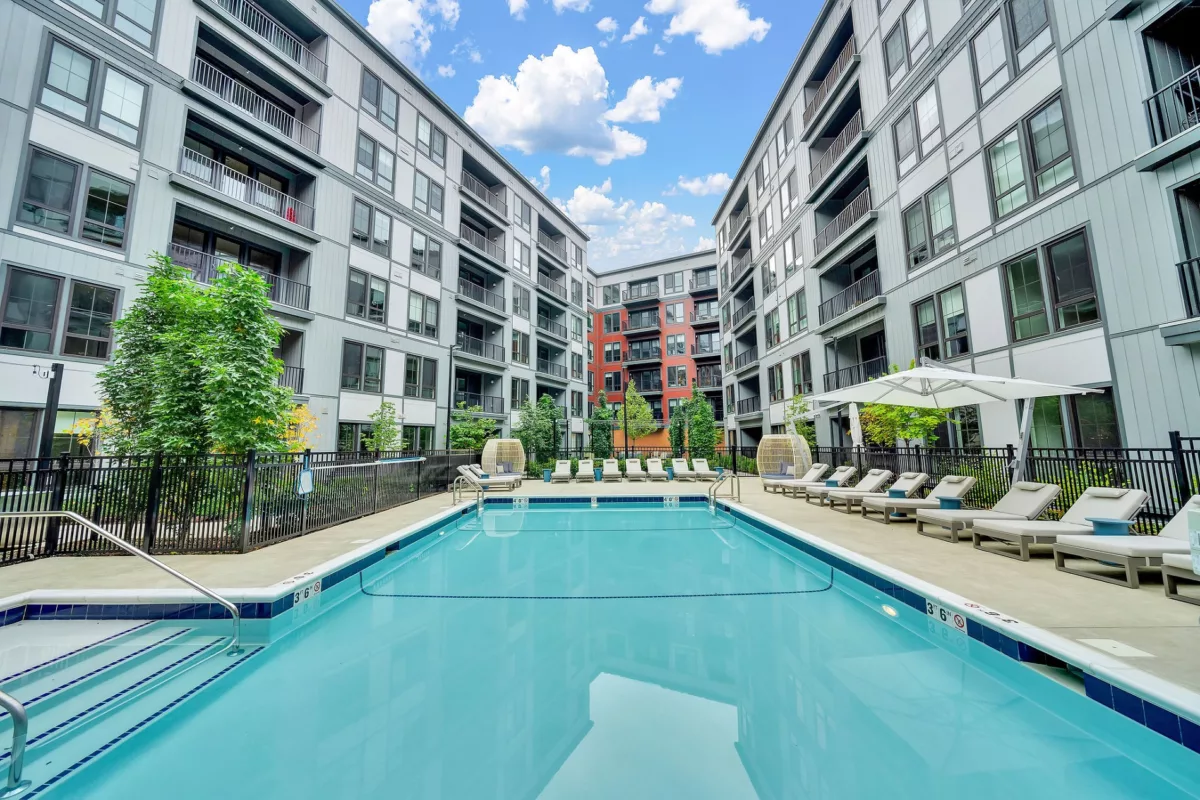Outdoor pool with lounge chairs surrounded by modern apartment buildings under a partly cloudy sky.