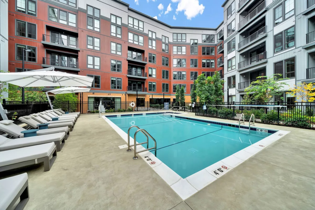 Outdoor pool with lounge chairs, umbrellas, and apartment building in the background.
