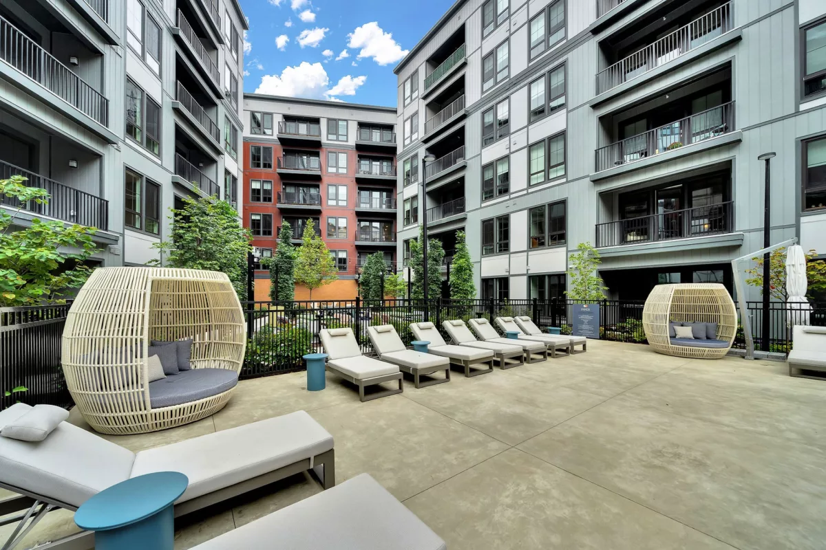 Outdoor lounge area with daybeds and wicker pod seating between modern apartment buildings.