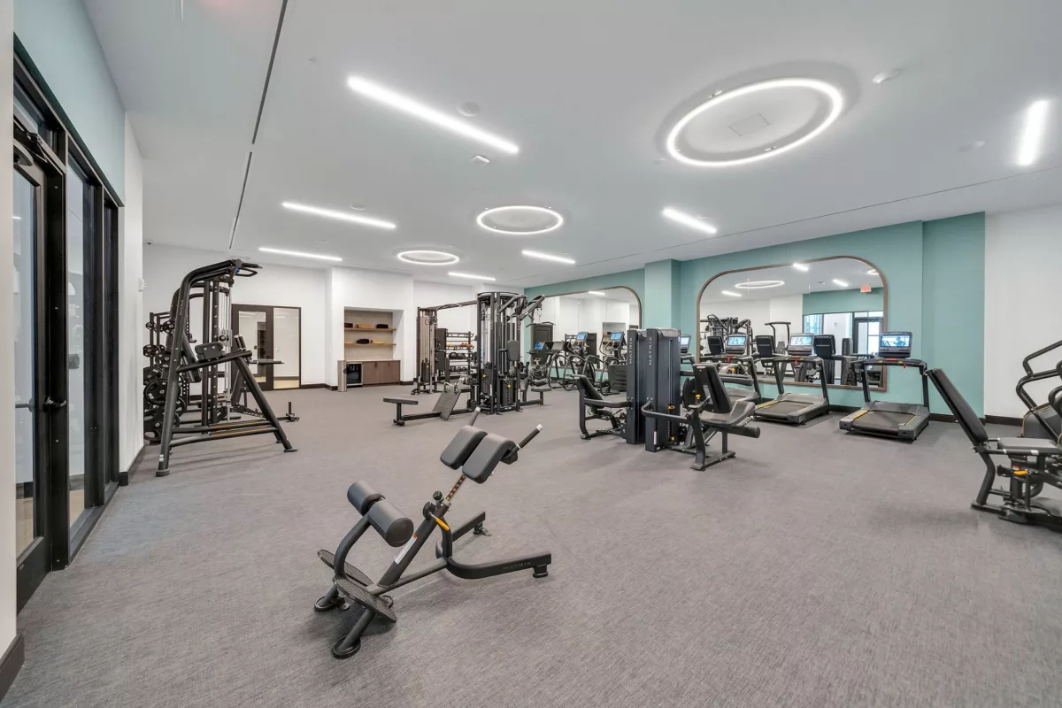 Fitness center with treadmills, weight machines, and mirrored wall. Gray carpet flooring.