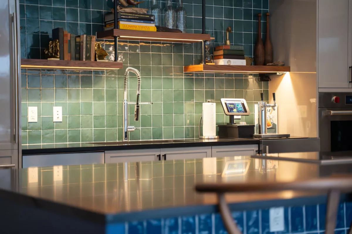 Kitchen with green tile backsplash, floating shelves, black countertops, and stainless steel faucet.