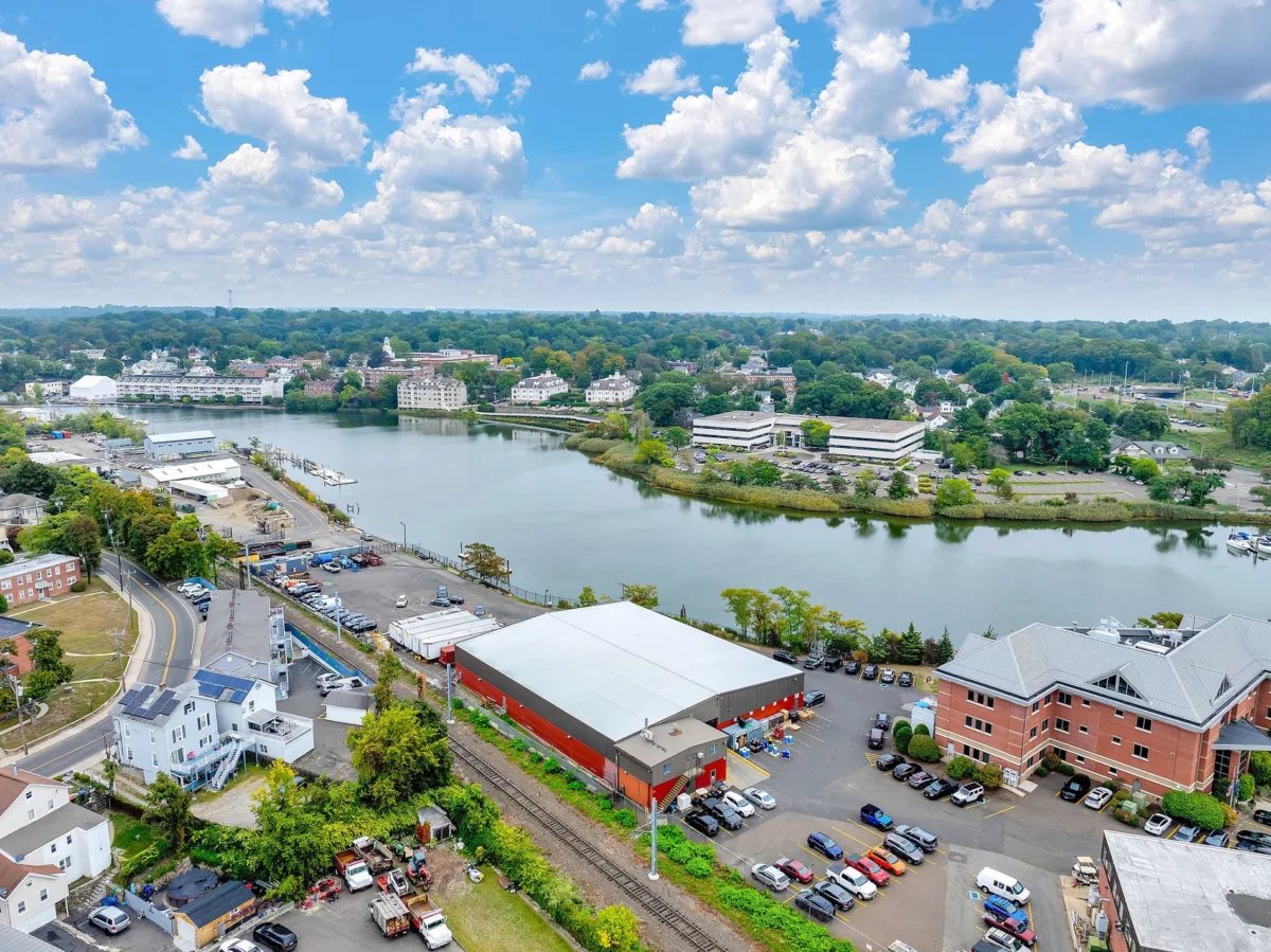 Aerial view of buildings, parking lots, and a river under a blue sky with scattered clouds.