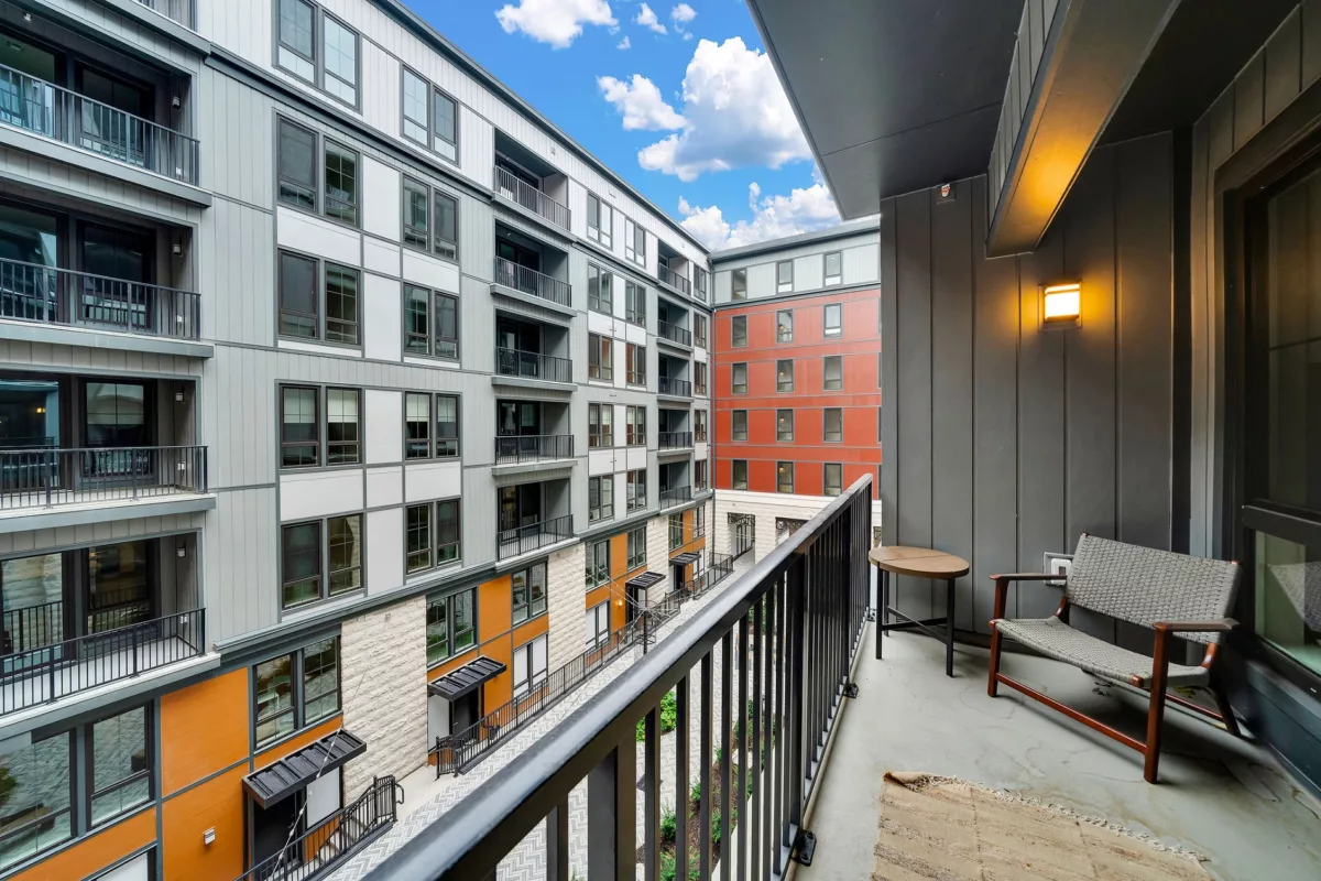Balcony with chair, table, and view of building courtyard under a blue sky with scattered clouds.