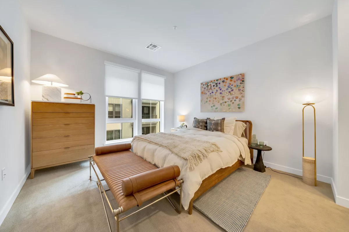 Bedroom with bed, dresser, leather bench, and modern lighting. Window with roller shades.