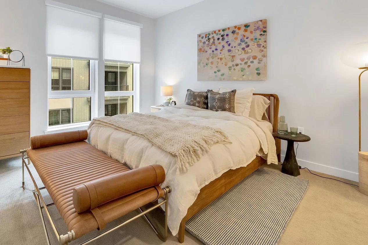 Bedroom with bed, dresser, leather bench, and modern lighting. Window with roller shades.