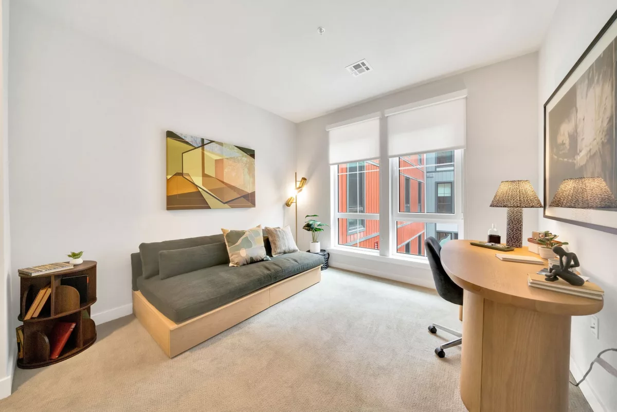 Bright room with daybed, desk, and large window showing exterior building. Neutral carpet.