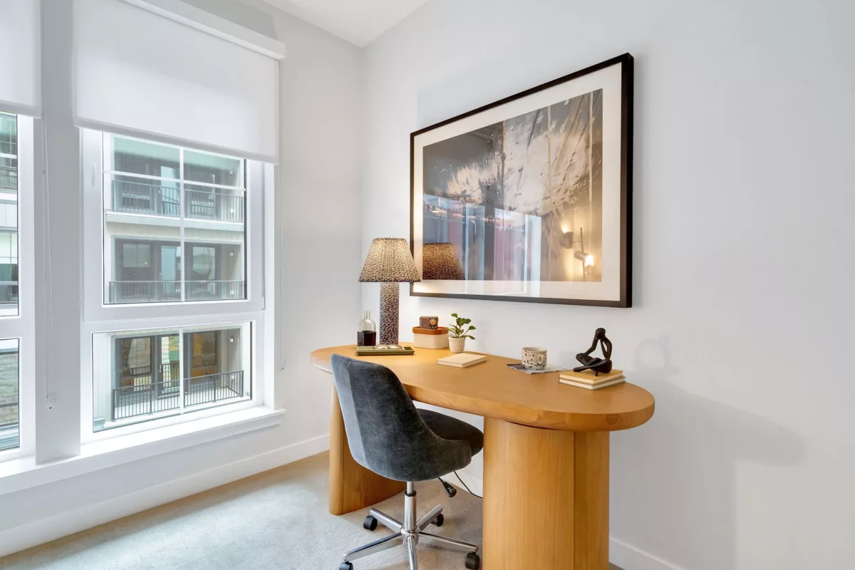 Interior view of a desk area with a wooden desk, chair, and window with white blinds.