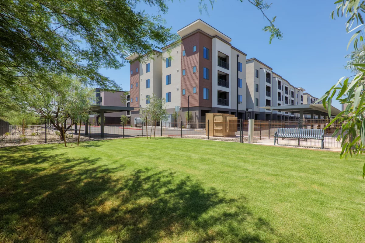Multi-story building exterior with green lawn, bench, covered parking, and desert landscaping.