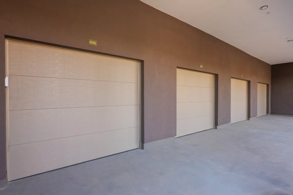 Row of beige storage unit doors along a brown wall with concrete floor and ceiling lights.