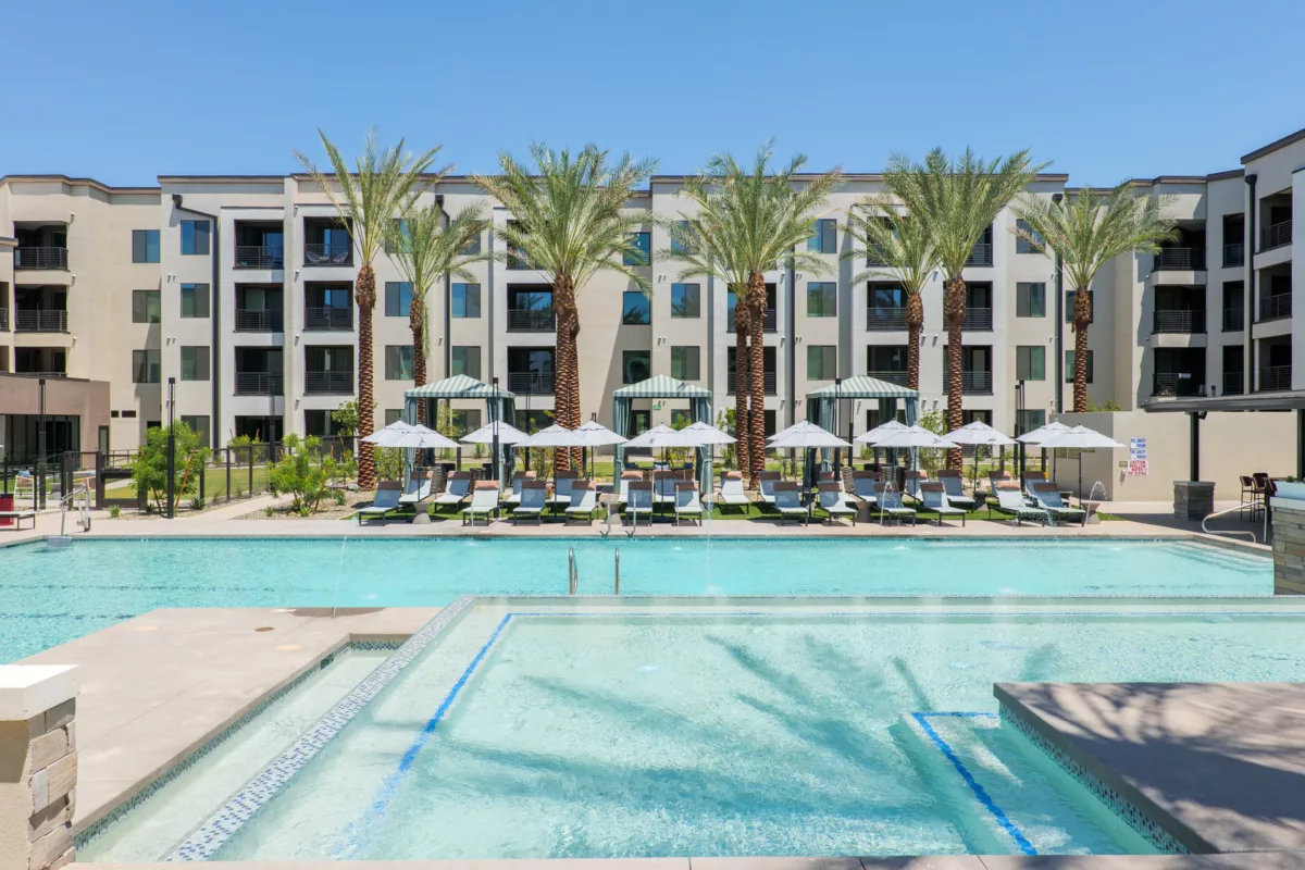 Outdoor pool with lounge chairs, umbrellas, and palm trees in front of a multi-story building.