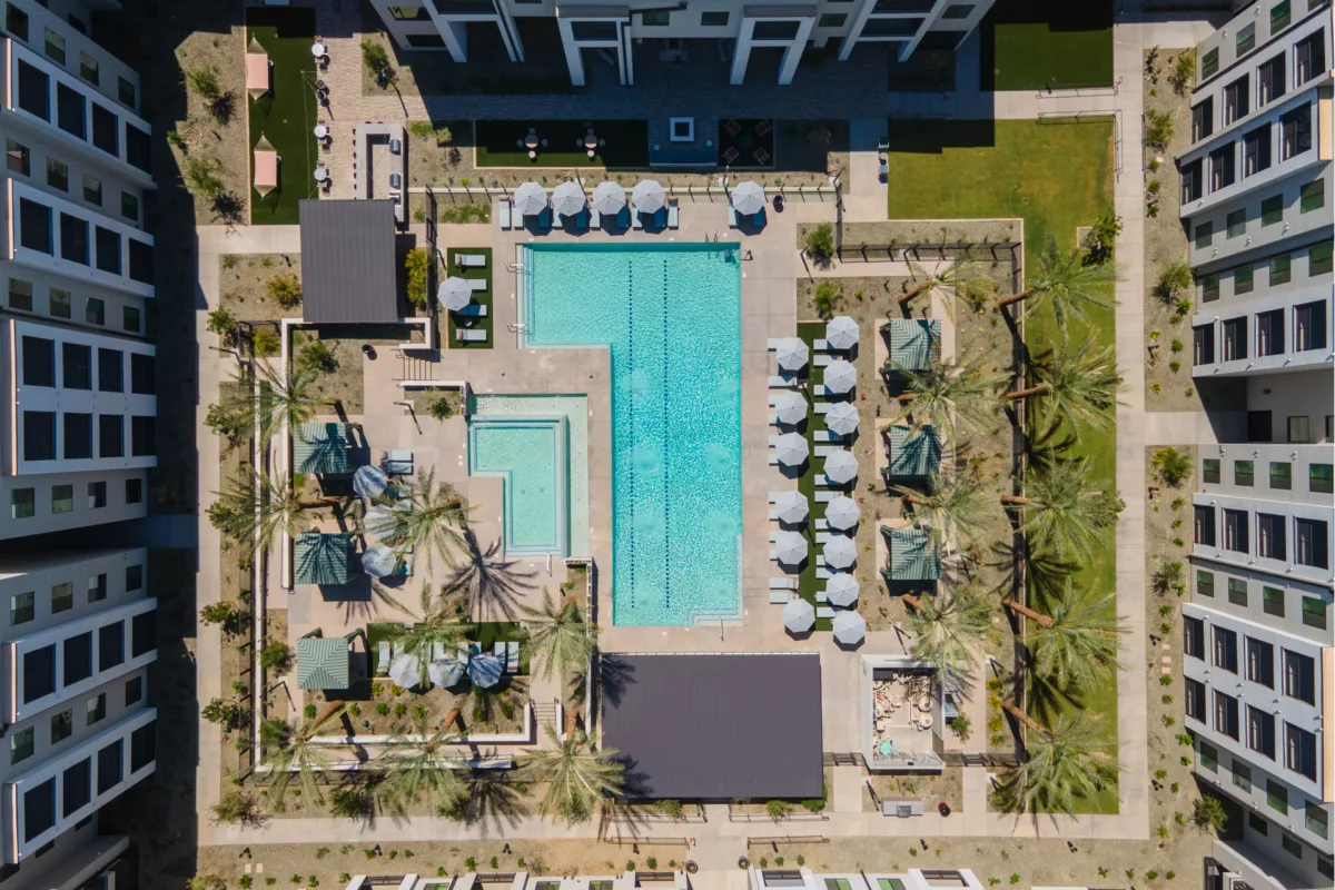 Aerial view of a pool, lounge chairs, umbrellas, and palm trees between apartment buildings.