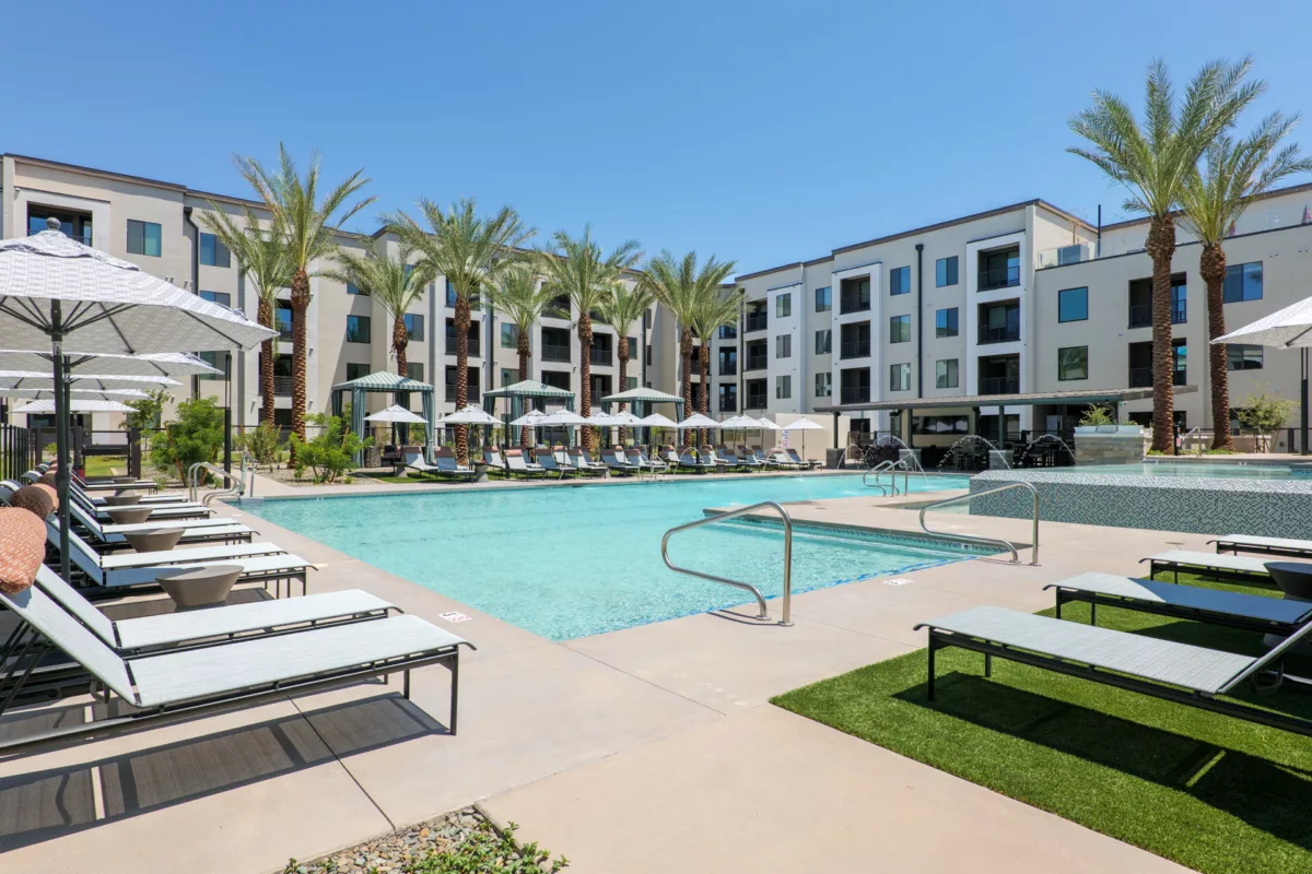 Outdoor pool with lounge chairs, umbrellas, and palm trees in front of a multi-story building.