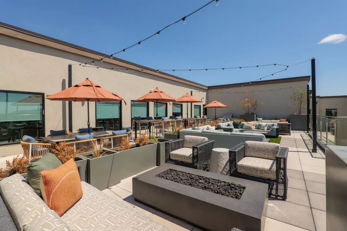 Rooftop lounge with seating, fire pit, dining tables, and string lights on a sunny day.