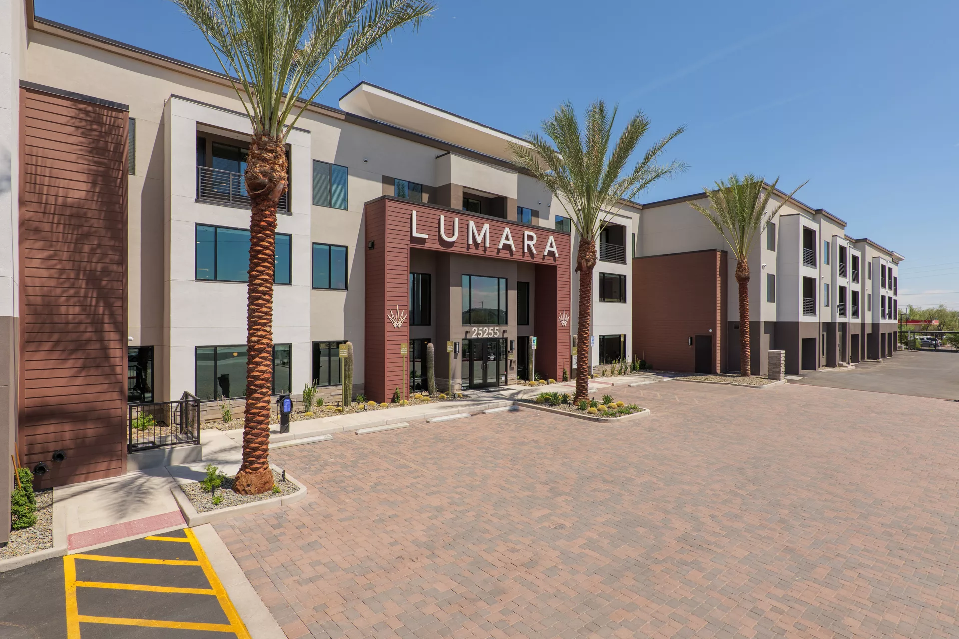 Exterior view of a modern apartment building with palm trees and brick paved parking area.