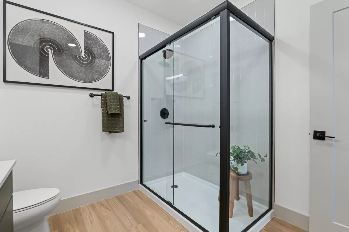 Modern bathroom with glass shower, black trim, light wood floors, and abstract art.