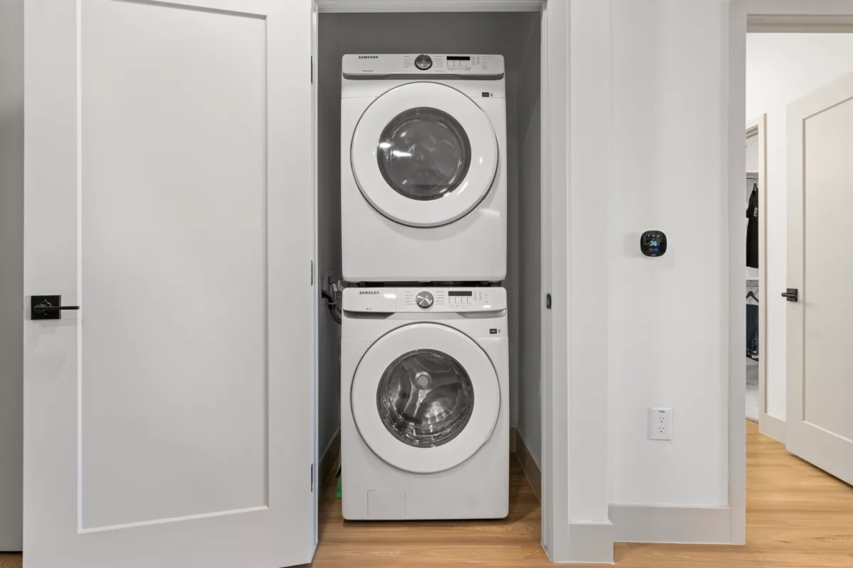 Stacked white washer and dryer inside a closet with an open white door and wood floors.