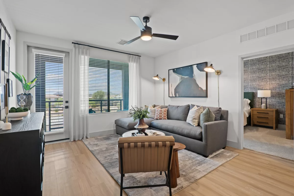 Modern living room with gray sofa, area rug, and view into a bedroom. Ceiling fan present.
