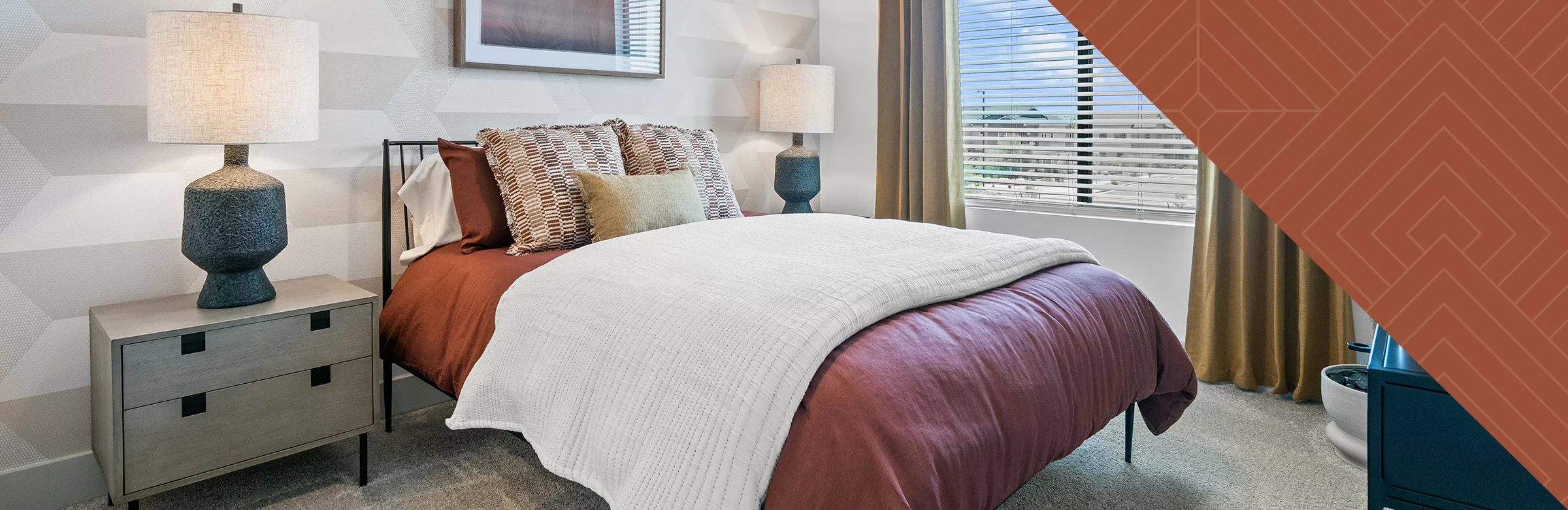 A photo of a bedroom with cream and red bedding and night stands with lamps in front of a wall with a geometric pattern.