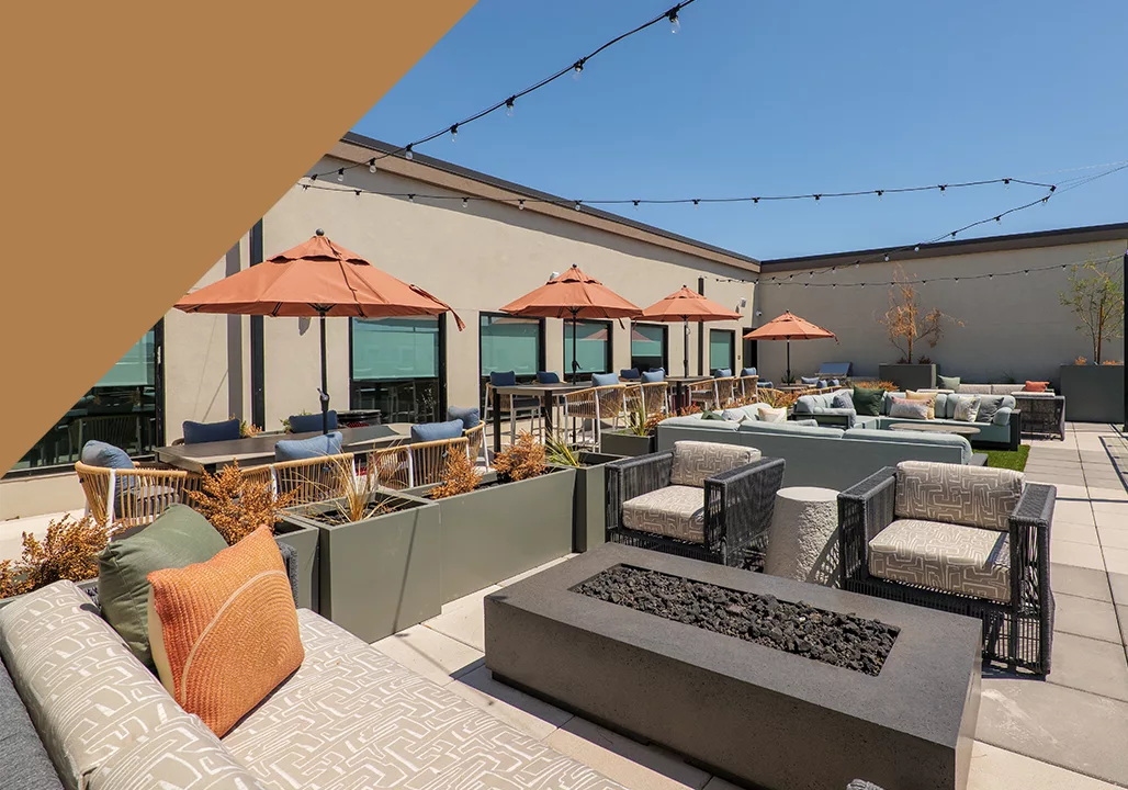 A photo of an outdoor lounge with seating about a fireplace and tables with umbrellas, with a decorative orange triangle in the upper left corner.