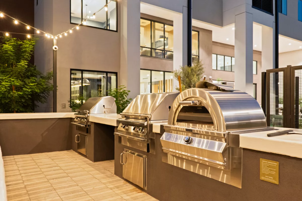 Outdoor grilling area with stainless steel grills and a pizza oven, illuminated by string lights.