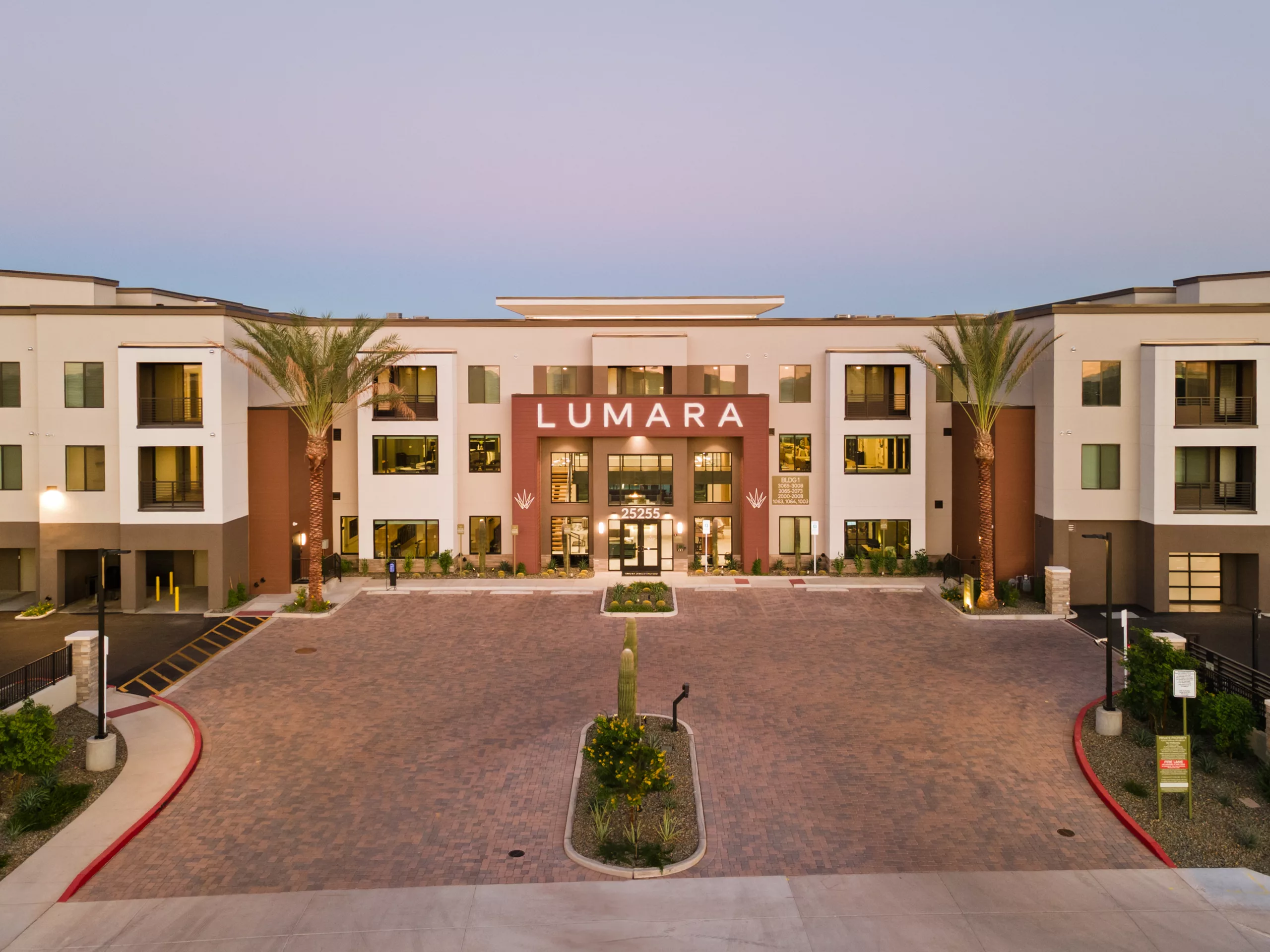 A building exterior with a brick driveway, palm trees, and a central entrance with glass doors under a sign that says "Lumara".