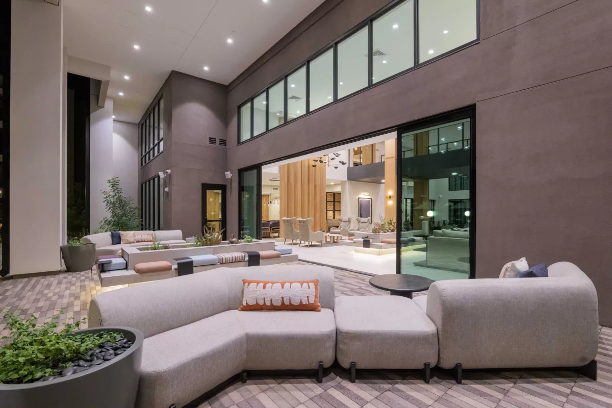 A modern lounge area features modular seating, planters, and large windows overlooking an adjacent seating area.