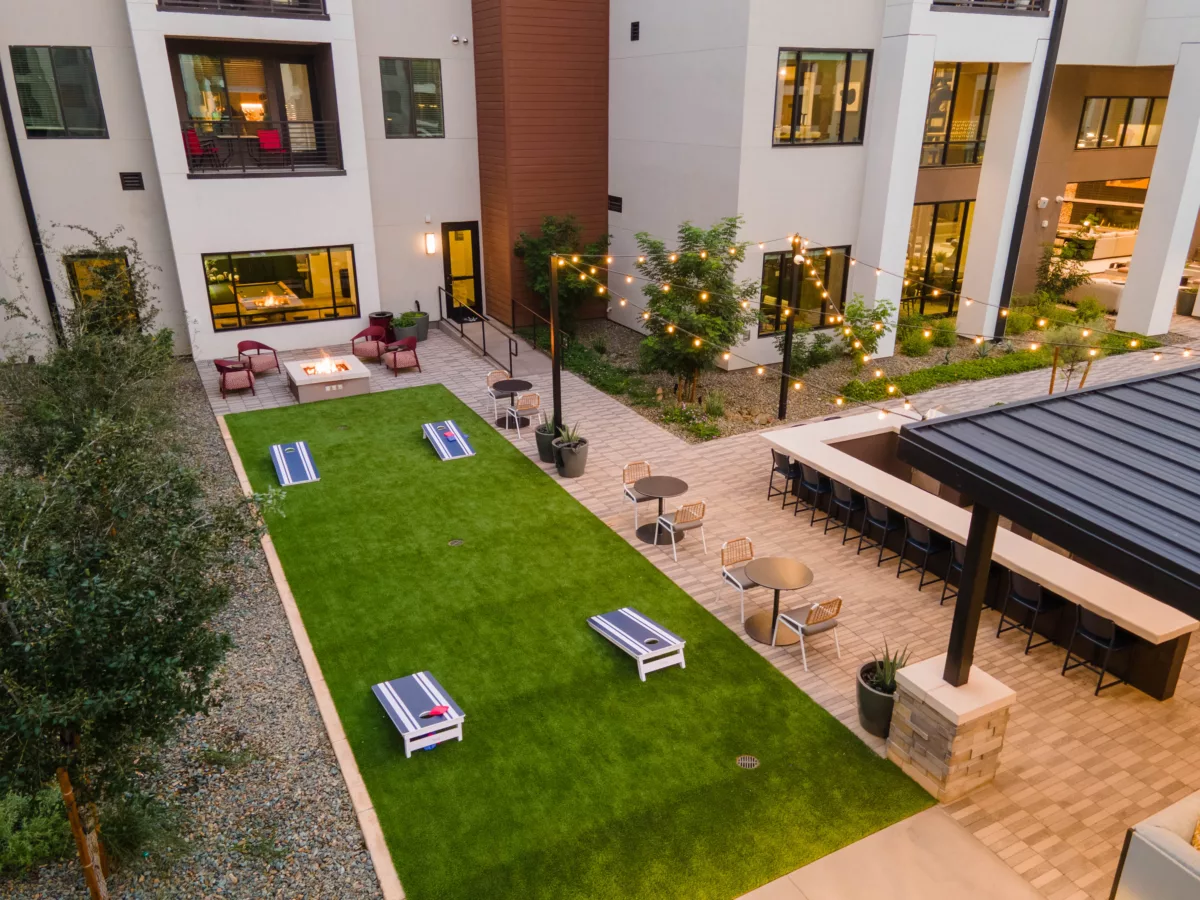 Aerial view of a courtyard with a cornhole game area, fire pit, outdoor bar, and string lights.