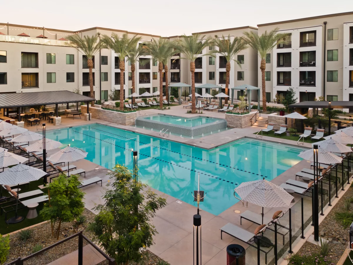 Outdoor pool area with lounge chairs, umbrellas, palm trees, and a covered seating area.