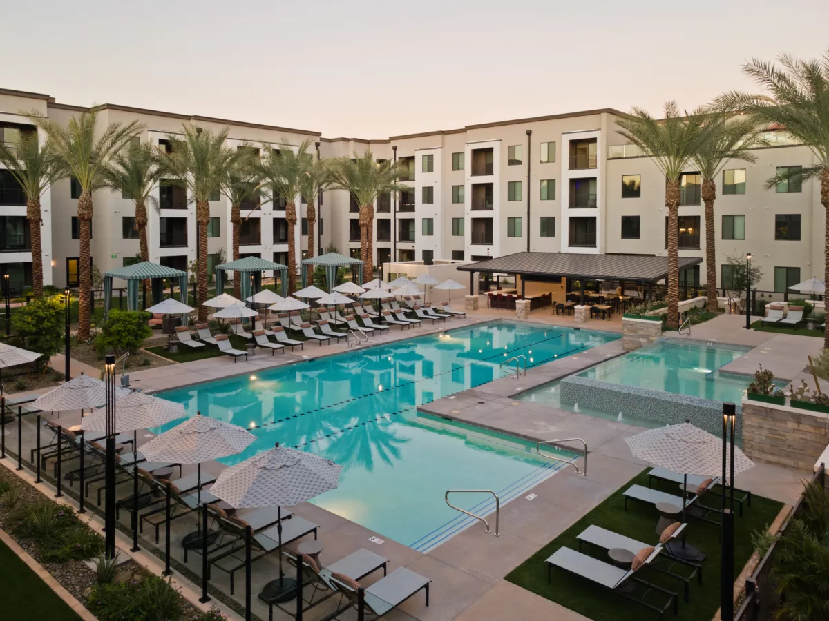 Outdoor pool area with lounge chairs, umbrellas, and a covered bar area in front of a multi-story building.