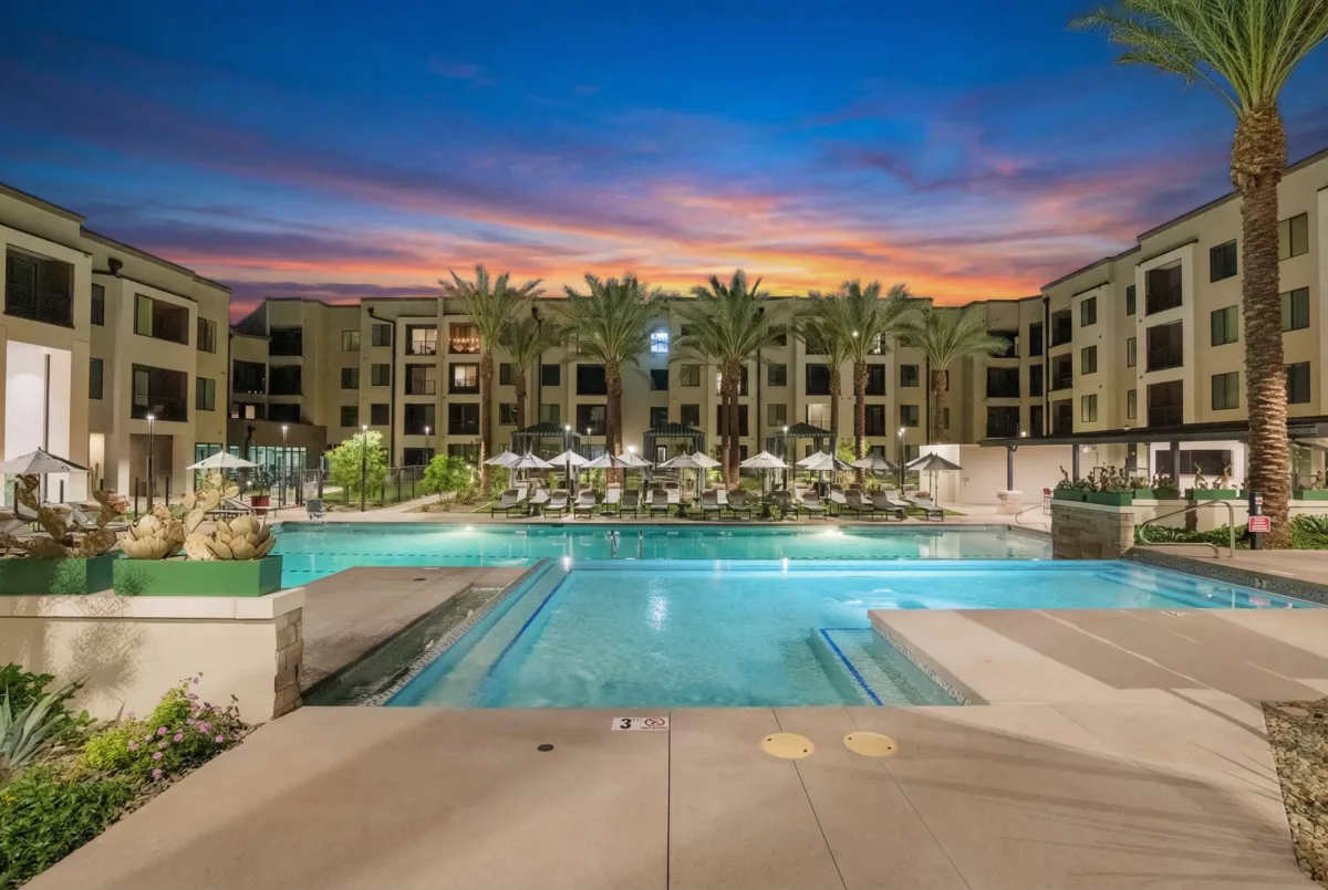 Outdoor pool area with lounge chairs, umbrellas, and palm trees between apartment buildings at dusk.