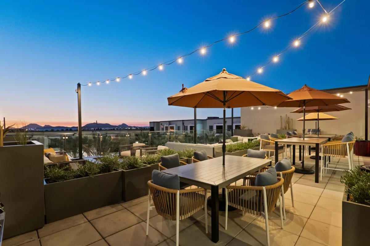 Rooftop lounge area features tables with umbrellas, cushioned seating, string lights, and mountain views at dusk.