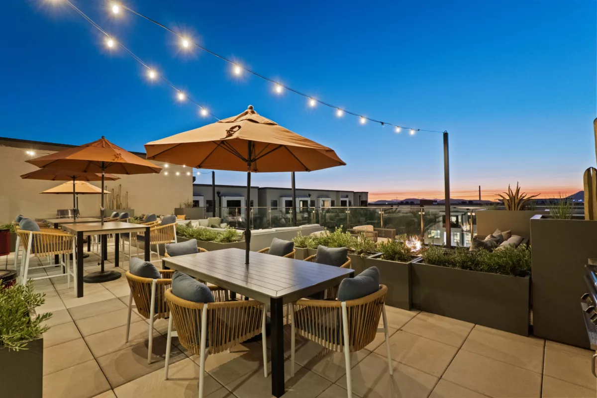 Rooftop lounge area with tables, chairs, umbrellas, planters, and a fire pit at dusk.