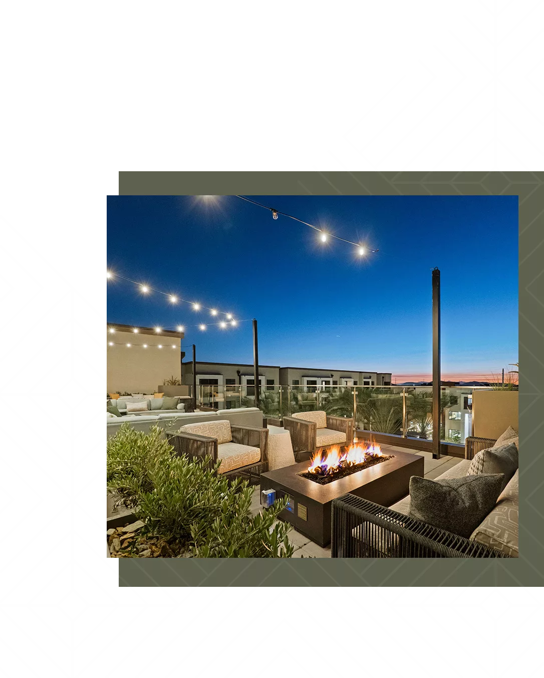 An outdoor rooftop lounge with armchairs, a fire pit, and string lights under a dusk sky.