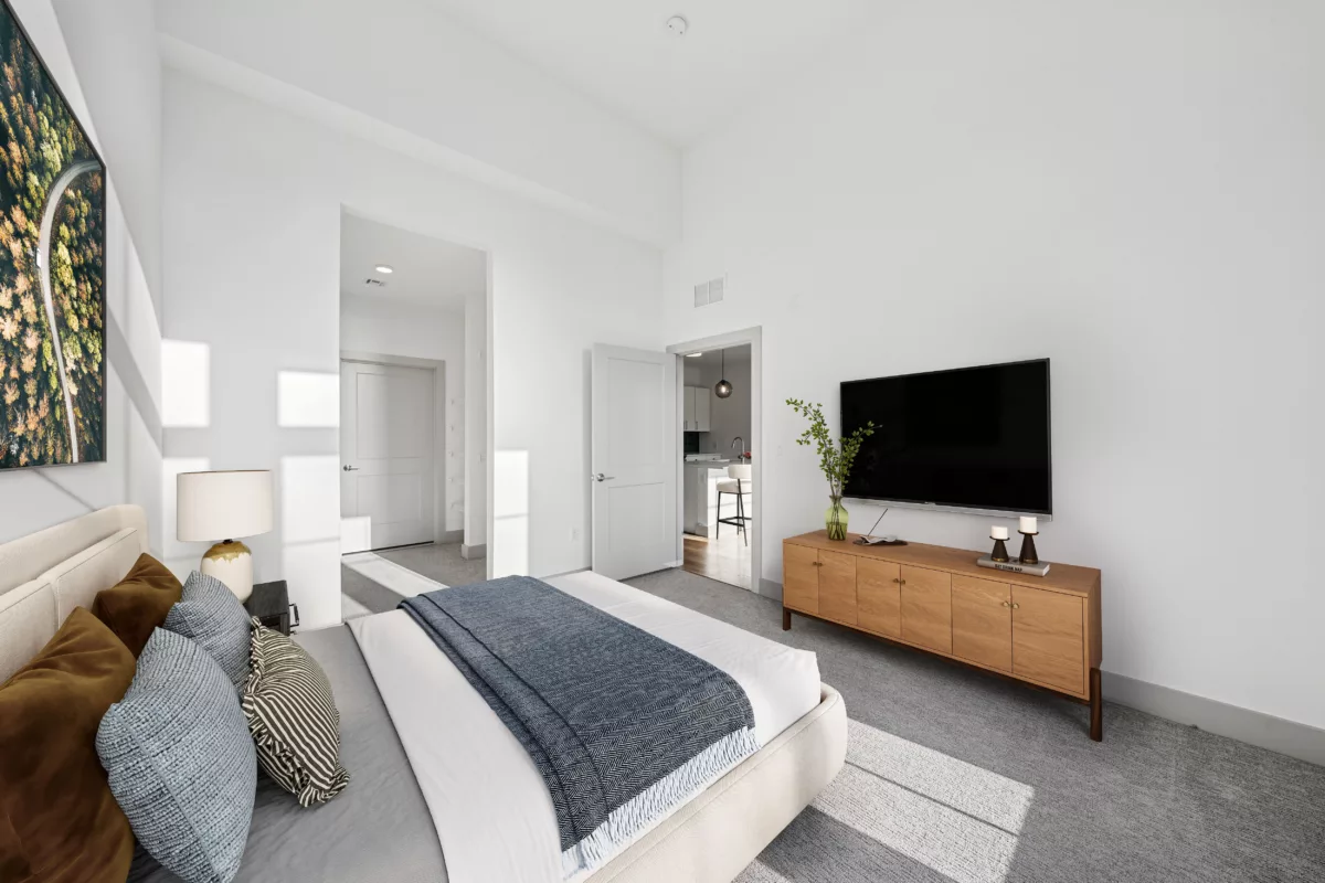 A bright bedroom with a bed, a wall-mounted TV over a wooden console, and a view into a kitchen.