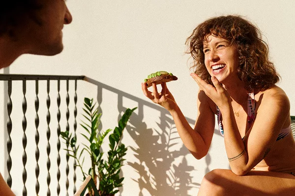 Two people outdoors, one holding avocado toast near railing and potted plant.