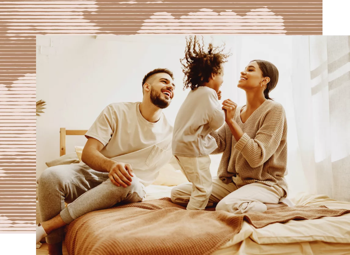 Smiling parents playing with toddler on a cozy bed in sunlit bedroom.