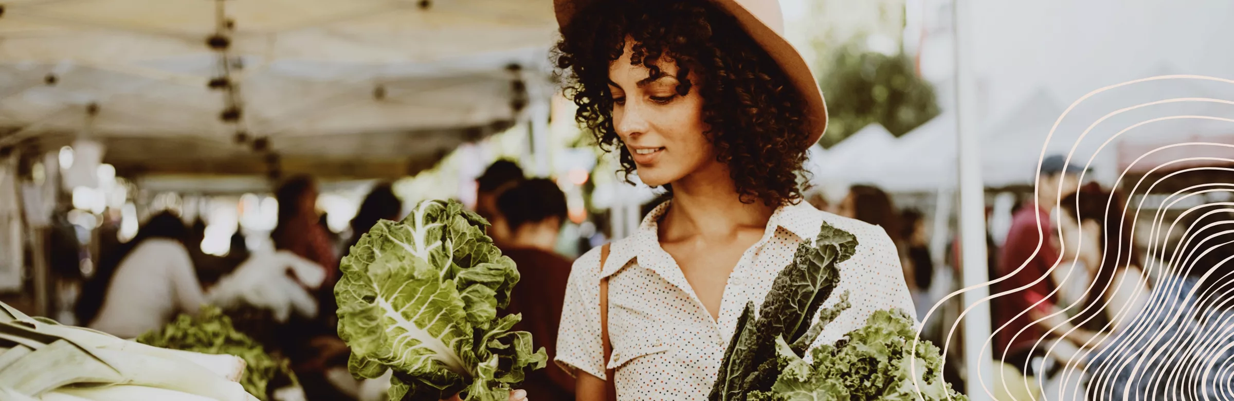 Woman shopping for leafy greens at an outdoor market under white canopy tents.