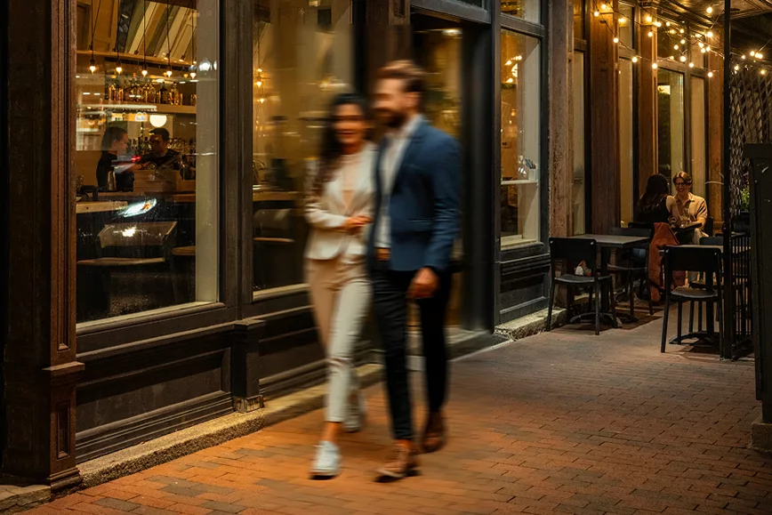 Blurred couple walking past lit restaurant with outdoor seating.