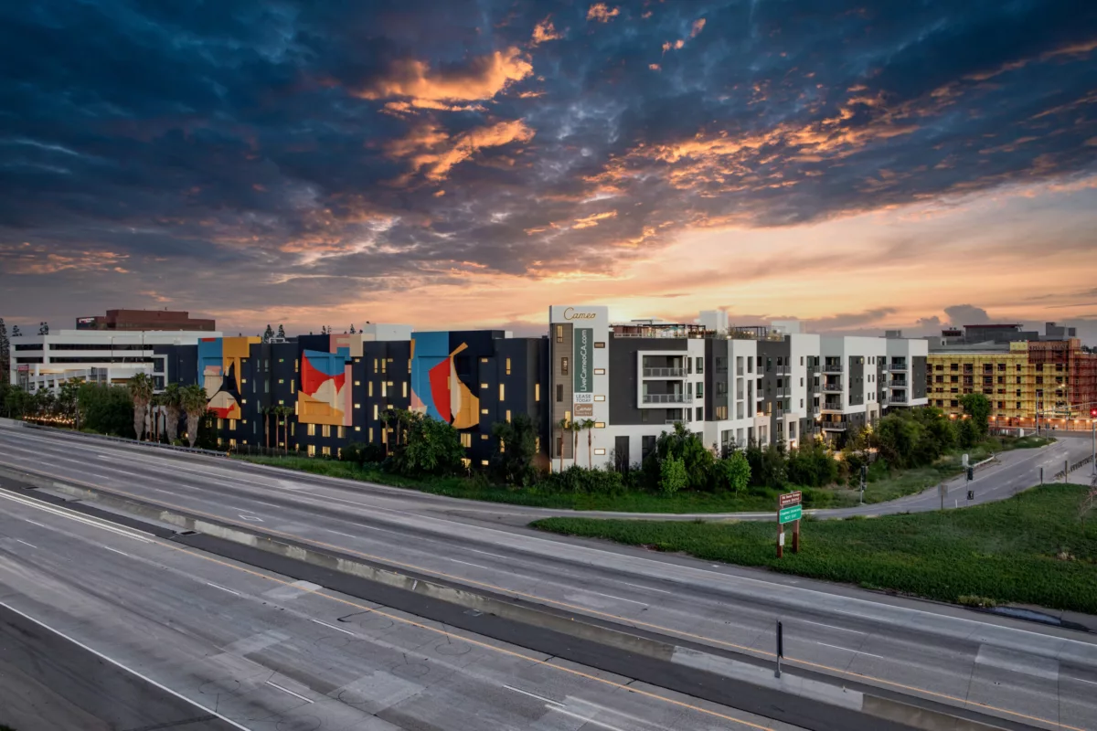 Artistic apartment complex beside a highway at sunset, featuring abstract mural-style designs on dark exterior walls and modern white-framed units.