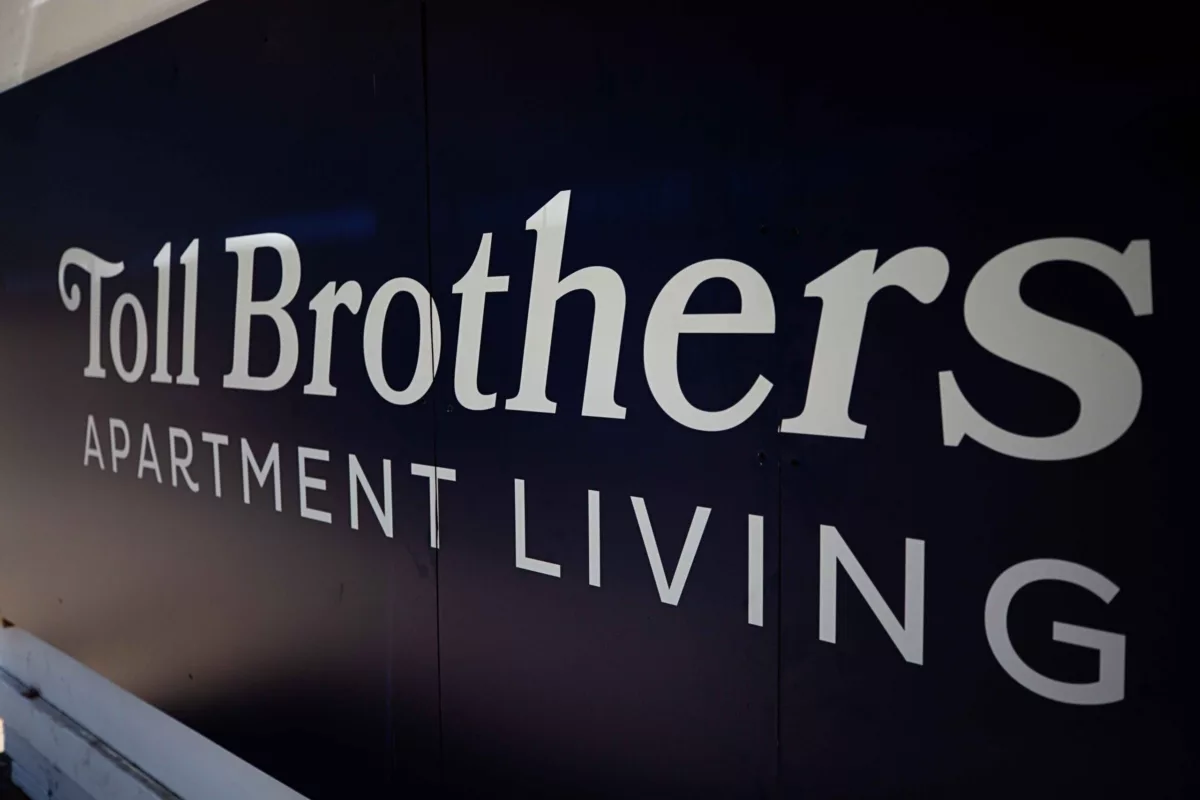 A close-up of a large sign that reads “Toll Brothers Apartment Living” in bold white letters on a dark background, representing the developer’s branding.