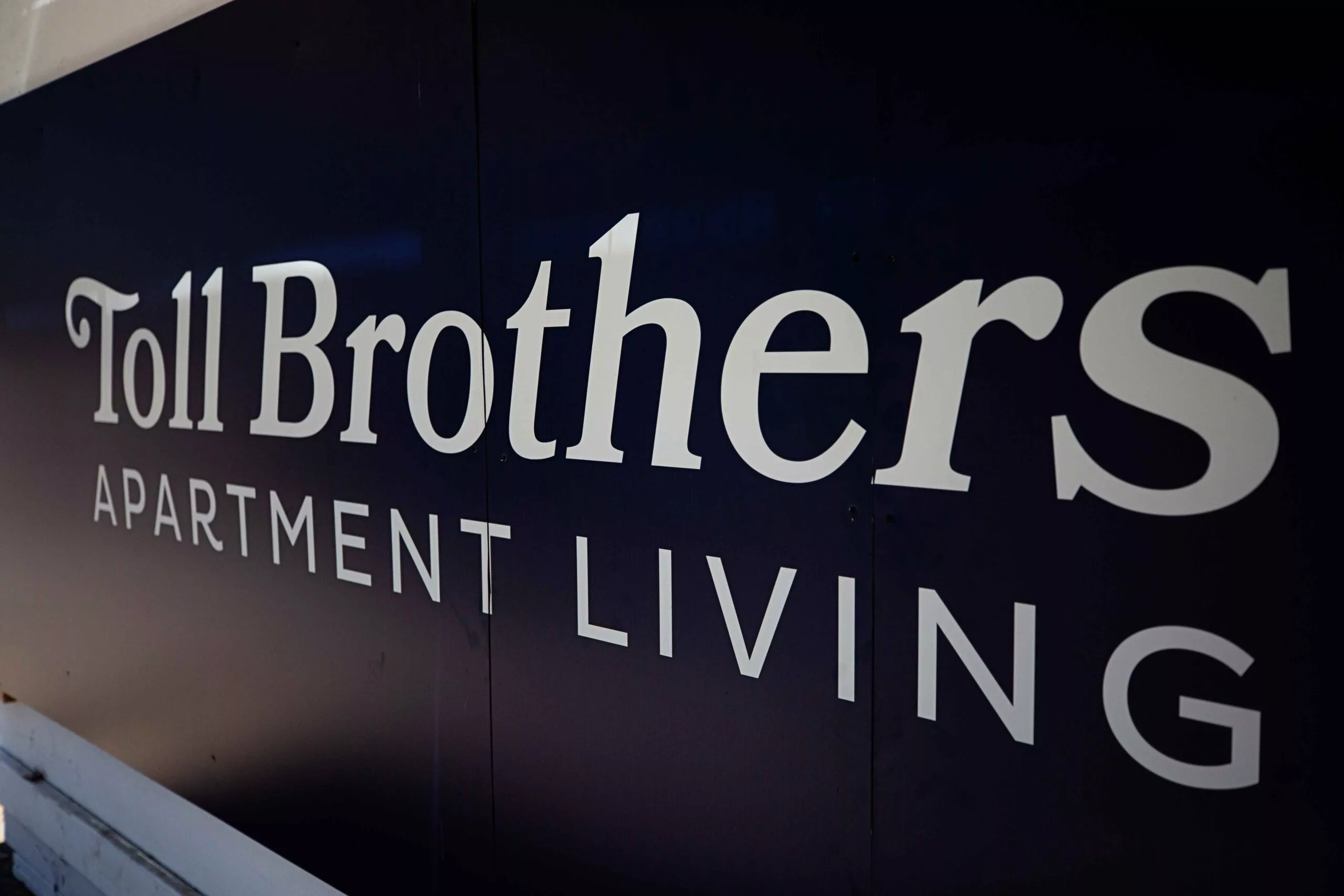 A close-up of a large sign that reads “Toll Brothers Apartment Living” in bold white letters on a dark background, representing the developer’s branding.