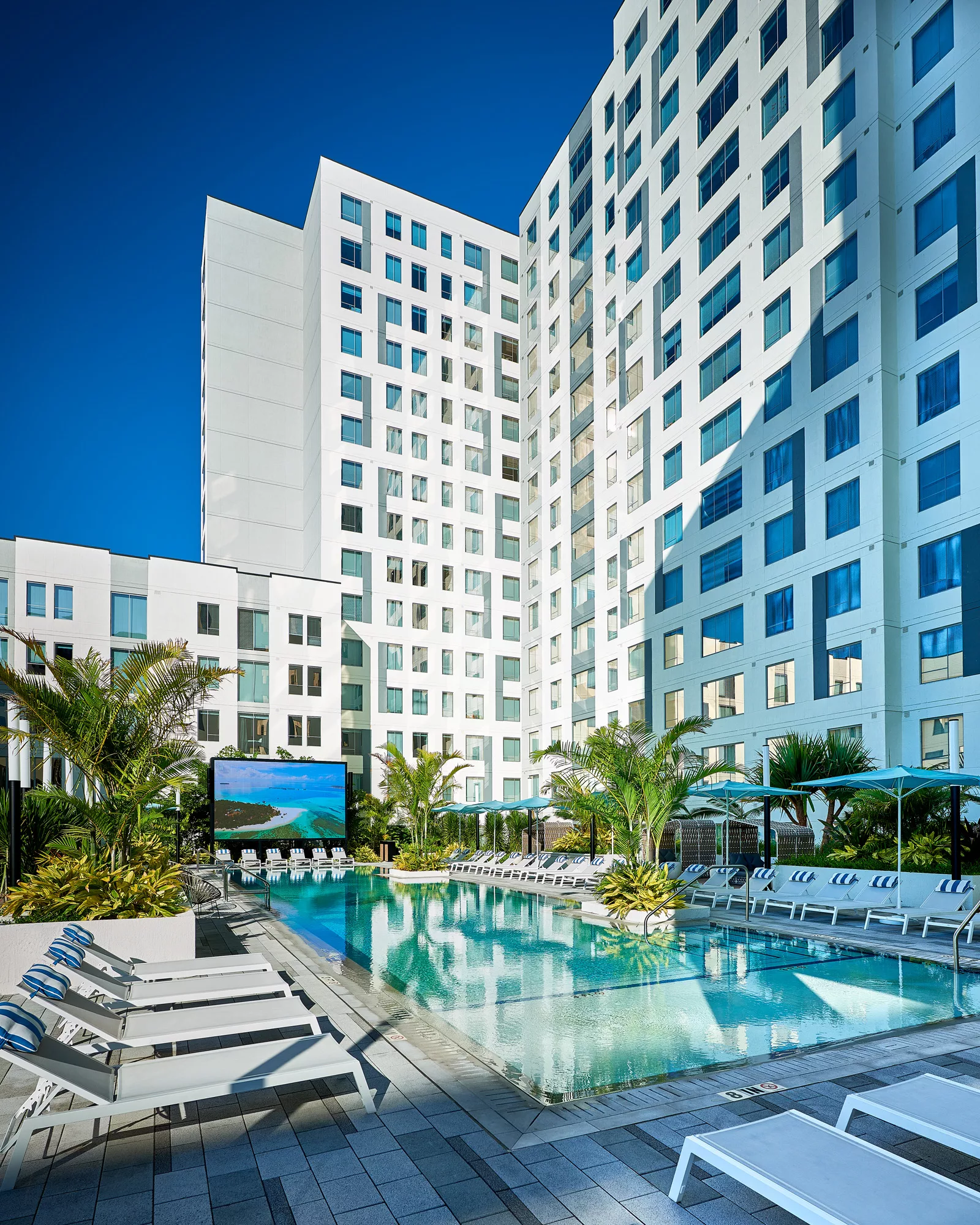 Pool area with tall buildings, palm trees, and a large outdoor screen